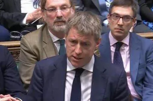 James, dressed in sober suit and tie, and with colleagues behind him,  makes a point in the House of Commons