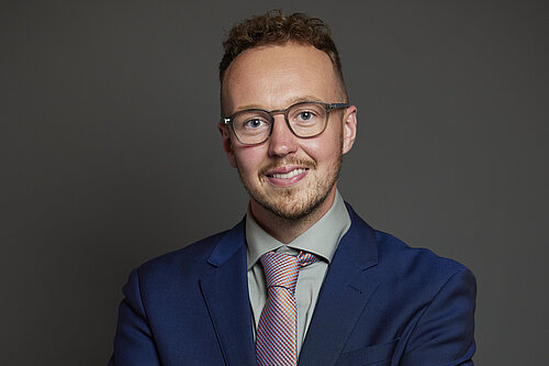 Adam Dance MP standing against a plain dark background, smiling with arms folded. He is wearing glasses, a blue suit, a light green shirt and a red and blue patterned tie.