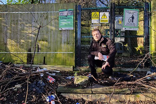 Councillor Charlie Clinton in front of a sign that says "Litterers, we're watching you", with a whole bunch of dumped litter