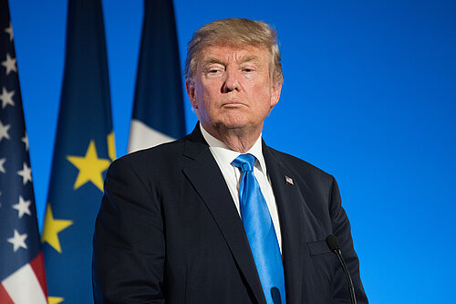 An image of Donald Trump standing in front of a blue backdrop with the US, EU, and French flags behind him