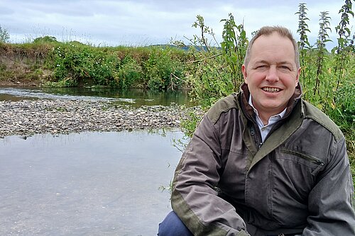 A photo of Richard Foord crouched down by a river. Behind him is a small island made of stones and some greenery.