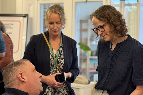 Alison Bennett MP speaks with attendees at a carers’ event, standing beside a seated man and another woman in a bright community room.