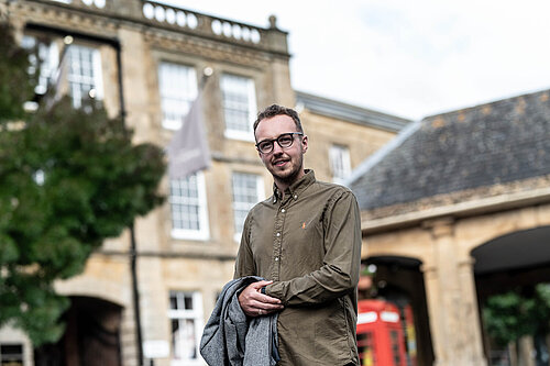 Adam Dance MP standing outside in front of a historic stone building, holding a grey coat and smiling at the camera.