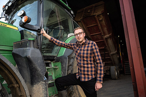 Adam Dance stands in a checkered shirt, with one arm on the door of a large, green tractor.
