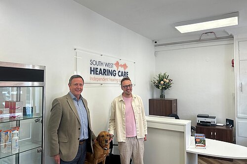 Two men and a guide dog stand in the reception area of South West Hearing Care beneath the company sign.
