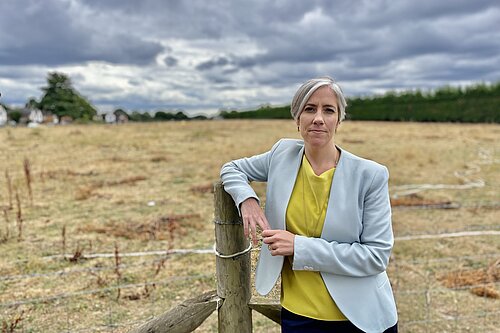 Daisy is leaning on a fence, standing in front of an area of green space and looking directly at the camera. 