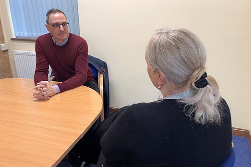 Ian sitting at a desk in a meeting room talking to someone else