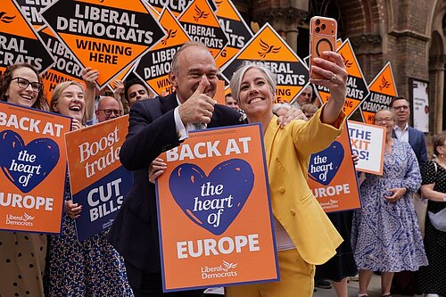 Ed Davey and Daisy Cooper take a selfie at a Liberal Democrat event, holding orange “Back at the heart of Europe” placards. Alison Bennett MP is visible at the far left, alongside supporters holding Liberal Democrat and “Winning Here” signs.