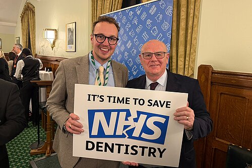 Two men stand indoors smiling at the camera while holding a sign that reads It’s time to save NHS dentistry.