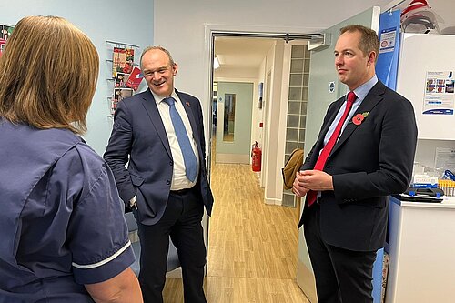 Ed Davey and Richard Foord standing in the corridor of a medical building, speaking to a nurse who has her back to the camera. Ed Davey is on the left in a suit with a bue tie, and Richard on the right in a suit with a red tie and a Remembrance poppy