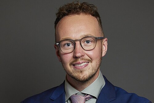 Adam Dance MP standing against a plain dark background, smiling with arms folded. He is wearing glasses, a blue suit, a light green shirt and a red and blue patterned tie.