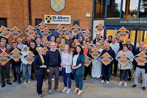 A group of Lib Dem councillors are standing in front of the St Albans District Council building, holding diamond shaped Liberal Democrat placards. Daisy Cooper MP, Councillor Paul de Kort, Councillor Helen Campbell and Victoria Collins MP (from left to right) are standing in front of the group.