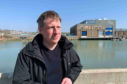 James in casual pose looking seriously into the distance with the river at Newhaven and industrial buildings behind him.