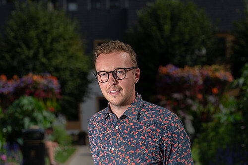 Adam Dance MP standing outdoors in a garden with colourful flowers and greenery, wearing a dark shirt with a red floral pattern and glasses.