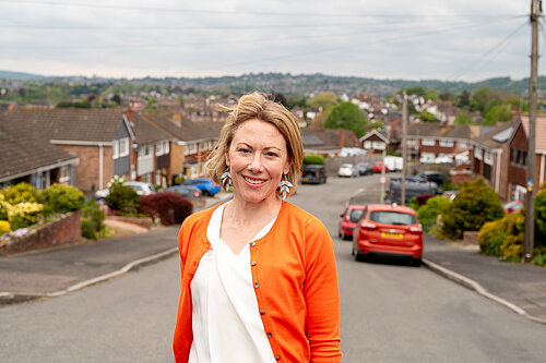 Charlotte FitzGerald in an orange cardigan and white shirt smiling while standing in a residential street