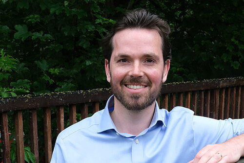 Adam Harley in a blue shirt standing with arm on top of a wooden fence of a bridge..