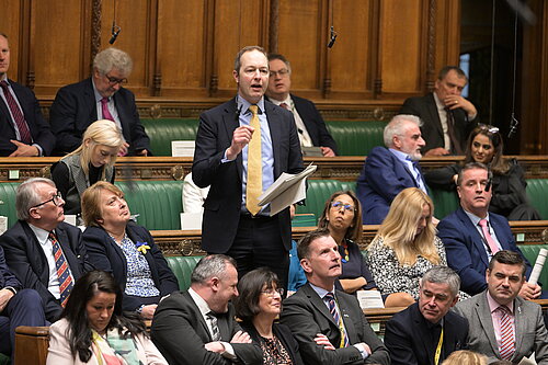 A photo of Richard Foord speaking in Parliament. He is surrounded by other MPs, and is wearing a black suit with a gold tie
