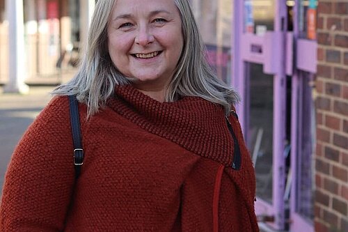 Councillor Lesley Boniface outside and looking cheerful. An entrance door behind her decorated in an uninviting mauve clashes with her dark red jumper.